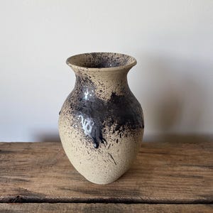 May include: A ceramic vase with a textured, beige, speckled surface featuring a dark brown, glossy glaze dripping from the rim down the upper body. The vase has a wide, flared opening and a rounded, bulbous shape that tapers towards the base.
