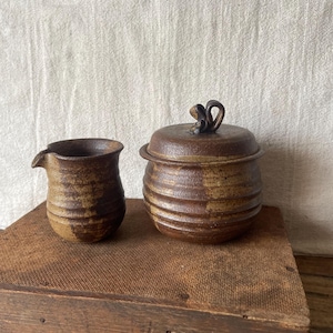 May include: A small, rustic ceramic creamer and lidded jar set. The creamer has a spout, and the jar has a loop handle on the lid. Both pieces are brown with a textured, earthy appearance, and sit on a wooden surface.