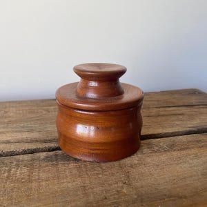 May include: A small, round, brown wooden container with a matching lid. The lid features a central, raised, circular knob handle. The body of the container has a smooth, cylindrical shape with a visible wood grain texture throughout the surface.