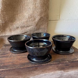 May include: A set of four dark, glossy, black ceramic bowls, each featuring a wide, flared rim and a matching circular base plate. The bowls have a smooth, reflective finish with subtle brown undertones visible along the edges and rims.