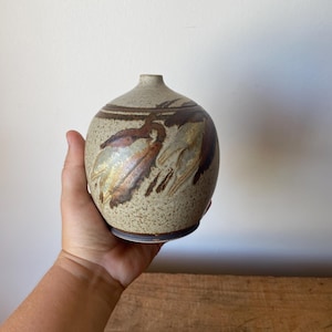 May include: A speckled, round ceramic vase with a narrow neck. The vase features brown and tan abstract leaf-like designs. The vase is held in a hand, showcasing its size and shape. The pottery has a rustic, handmade aesthetic.