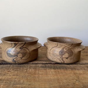 May include: Two round ceramic bowls with small side handles, featuring a speckled tan base colour and dark brown swirling marbled patterns. The bowls have a wide opening and a smooth, matte-textured finish, showcasing a consistent earthy design.