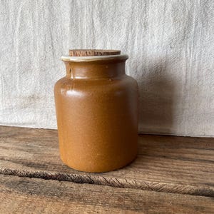 May include: A small, cylindrical brown ceramic jar with a smooth, matte finish and a slightly flared rim. The jar is topped with a circular, flat-topped cork stopper that fits snugly into the opening, providing a simple and functional closure.
