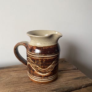May include: A ceramic pitcher featuring a two-tone glaze with a speckled cream-coloured rim and a dark brown body. The lower section of the pitcher is decorated with horizontal, wavy, incised lines that reveal the lighter clay underneath the glaze.