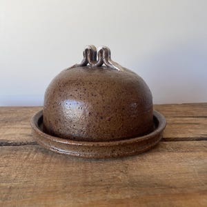 May include: A brown ceramic butter dish with a speckled glaze finish. The dome-shaped lid features a small, looped handle at the top, and the base consists of a circular plate with a raised rim to secure the lid in place during storage.