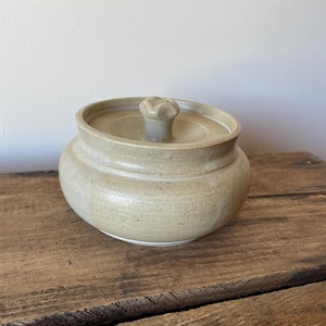 May include: A round, beige ceramic lidded jar with a smooth, speckled glaze finish. The jar features a wide, curved body and a small, sculpted knob handle on the centre of the lid. The ceramic surface displays subtle variations in tone and texture.