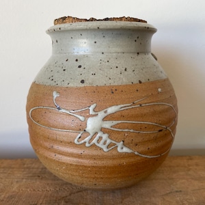 May include: A handcrafted ceramic jar with a cork stopper. The jar features a speckled, light grey top and a tan body with white abstract designs. The jar is approximately 15 cm tall.