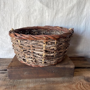 May include: A round, open-top basket constructed from interwoven brown twigs and bark strips. The basket features a thick, braided rim made of darker brown wood, with a textured, rustic exterior showing natural variations in wood colour and thickness.