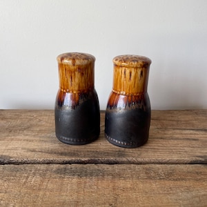 May include: A pair of ceramic salt and pepper shakers featuring a cylindrical shape with a rounded top. The upper portion has a glossy tan and brown drip glaze, while the lower portion is finished in a matte black with a beaded base detail.