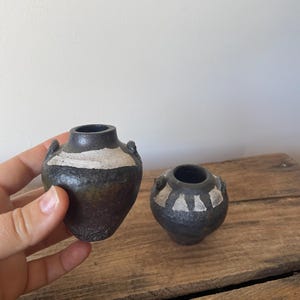 May include: Two small, dark, textured ceramic vases with narrow openings and side handles. The larger vase features a wide, horizontal white band, while the smaller vase is decorated with a series of white triangular patterns around the neck.