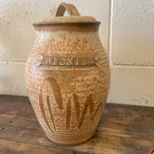 May include: A tall, cylindrical ceramic jar with a matching lid and a handle on top. The jar features a textured, speckled beige and tan glaze with a decorative banner across the front containing the embossed text RICKIES in capital letters.