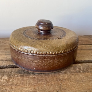 May include: A round, lidded ceramic container featuring a two-tone brown glaze. The base is a deep reddish-brown, while the lid is a lighter tan with a central knob handle and a decorative embossed beaded border circling the top edge.