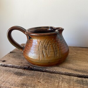 May include: A small, round ceramic creamer jug featuring a glossy, variegated brown and amber glaze. The exterior surface displays a series of vertical, incised linear markings around the body, complemented by a curved handle and a small spout.