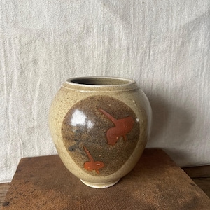 May include: A round, beige ceramic vase with a dark brown circular design featuring two orange abstract shapes. The vase has a wide opening and sits on a wooden surface. The background is a light-coloured fabric.