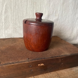 May include: A small, cylindrical dark brown wooden jar with a matching lid. The lid features a central knob handle and a small notch cut into the side, allowing for a spoon to rest inside while the lid is closed. The wood grain is visible throughout.