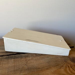 May include: A rectangular wooden display stand with a slanted top surface painted in an off-white colour. The structure features a flat base and a sloped top panel secured with visible metal screws at the corners, showing a distressed finish.