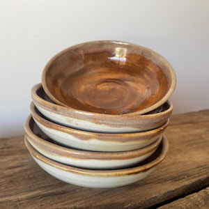 May include: A stack of four round ceramic bowls with a glossy brown interior glaze and a matte off-white exterior. The bowls feature a visible drip pattern where the brown glaze meets the off-white base, creating a layered, organic appearance.