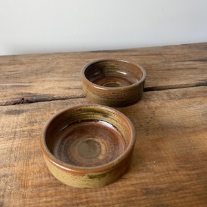 May include: Two round ceramic bowls with a low profile and straight sides. The bowls feature a variegated glaze in earthy tones of brown, tan, and ochre, with a smooth, glossy interior finish and a slightly textured, matte exterior surface.