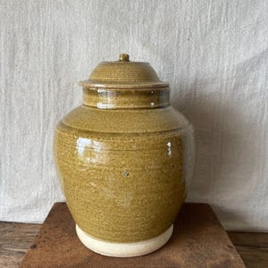 May include: A glazed ceramic jar with a lid. The jar is a muted olive green colour and sits on a white base. The lid has a small knob on top. The jar is on a wooden surface.
