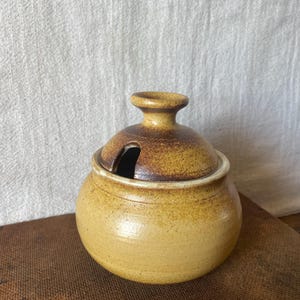 May include: A small, round, ceramic sugar bowl with a lid. The bowl is a light yellow colour with darker brown accents on the lid and rim. The lid has a small handle and a slot for a spoon. The bowl is sitting on a brown surface.