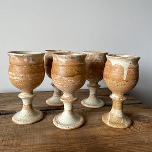 May include: A set of five ceramic goblets featuring a speckled tan and cream glaze with a rounded bowl, a flared rim, and a pedestal base. The pottery displays a natural, earthy colour palette with visible drip patterns and a textured finish.
