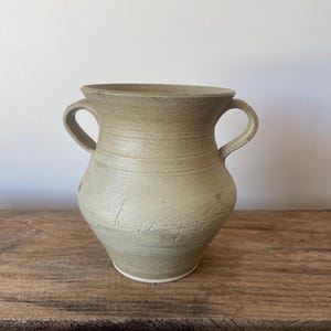 May include: A light beige ceramic vase with two side handles, a wide flared rim, and a rounded body. The surface features horizontal incised lines around the neck and subtle, diagonal etched markings across the midsection of the vessel.