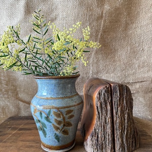May include: A ceramic vase with a speckled light blue glaze and painted brown and green leaf motifs, containing a sprig of yellow mimosa flowers, positioned next to two matching bookends made from natural wood logs with rough bark edges.