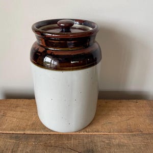 May include: A ceramic jar with a lid. The jar is mostly white with brown accents around the top and lid. The lid has a small, round knob. The jar is sitting on a wooden surface.