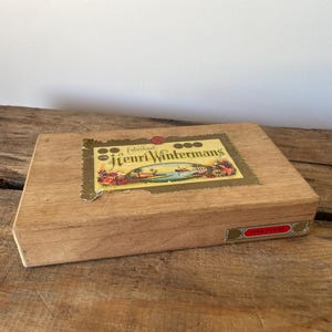 May include: A rectangular wooden cigar box with a light brown finish. The lid features a central label with the text Henri Wintermans, depicting a tropical landscape with a ship, palm trees, and colourful flowers. A small red label is on the side.