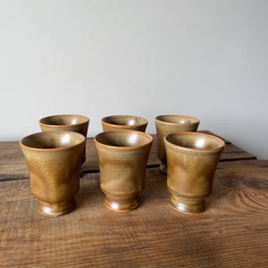 May include: A set of six ceramic cups with a speckled, earthy brown glaze. Each cup features a flared rim, a slightly tapered body, and a raised pedestal base. The cups are arranged in two rows, showcasing a consistent, textured finish throughout.