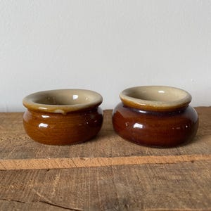 May include: Two small, round ceramic pots with a glossy brown exterior and a cream-coloured rim. The pots are on a rough, wooden surface, against a plain white wall.