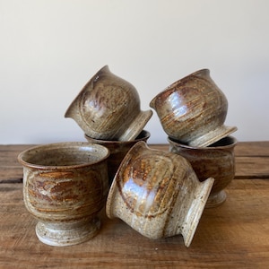 May include: A set of five ceramic cups with a speckled brown and tan glaze. Each cup features a rounded body, a flared rim, and a pedestal base. The glaze displays variations in colour intensity, with darker brown bands and lighter tan areas.