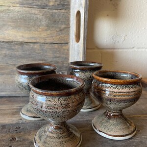 May include: A set of four ceramic egg cups featuring a speckled glaze in shades of tan, brown, and grey. Each cup has a rounded bowl, a flared pedestal base, and a smooth, glossy finish with a darker brown rim around the top opening.