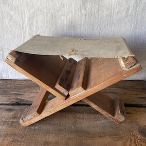May include: A vintage wooden footstool with an X-frame base and a woven fabric seat. The wood is a warm, natural colour, and the fabric is a light beige. The stool has a handmade appearance and a rustic aesthetic.
