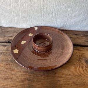 May include: A brown ceramic serving platter with a central dip bowl. The platter has a decorative band with white floral accents. The dish has a glossy finish and is set on a wooden surface.