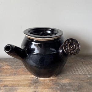 May include: A glossy black ceramic teapot with a side handle and a short spout. The circular handle features embossed Japanese characters arranged in a ring around a central symbol. The teapot has a wide, flat lid and a rounded, bulbous body.