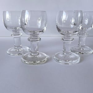 May include: A set of four clear glass stemmed glasses, each featuring a rounded bowl, a thick central stem with a prominent horizontal ring detail, and a wide, flat circular base. The glassware is transparent and uniform in design.