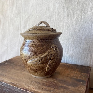 May include: A handmade, brown ceramic jar with a lid. The jar has a textured surface and is decorated with an embossed feather design. The lid has a loop handle. The jar is on a wooden surface.