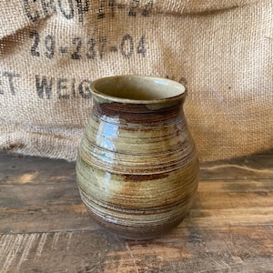 May include: A rounded ceramic vase featuring a variegated glaze in shades of earthy brown, tan, and olive green. The vessel has a wide, slightly flared rim and a smooth, bulbous body with subtle horizontal ridges circling the exterior surface.