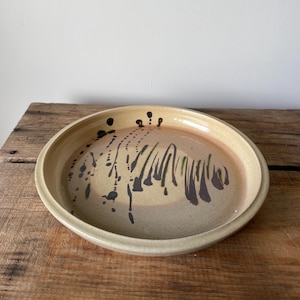 May include: A round ceramic plate with a raised rim, featuring a light beige base glaze. The surface is decorated with dark brown and black abstract drip patterns and vertical brush strokes that create a textured, organic visual effect.