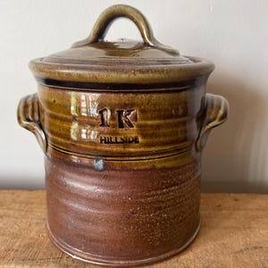 May include: A brown ceramic jar with a lid and handle. The jar has two side handles and the text "1K HILLSIDE" is visible on the side. The jar is sitting on a wooden surface.