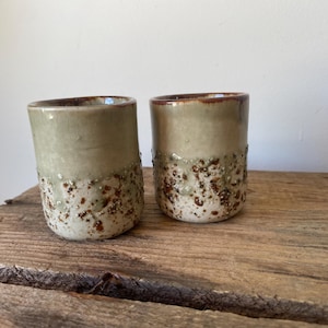 May include: Two cylindrical ceramic cups featuring a smooth, light sage green glaze on the upper portion and a textured, speckled cream and brown finish on the lower half, with a visible drip effect where the two distinct glaze textures meet.