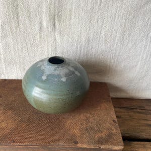 May include: A spherical ceramic vase with a small opening. The vase features a colour gradient, shifting from muted green at the base to blue-grey at the top. The vase is placed on a brown surface.