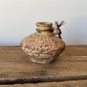 May include: A small, round, tan-coloured ceramic vessel with a textured, ridged surface and a weathered, mottled finish. A thin, dark brown rope is tied around the neck, featuring a single light-coloured wooden bead and a small, frayed tassel.