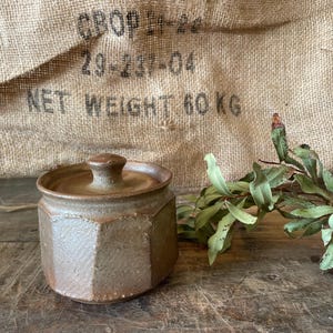 May include: A hexagonal ceramic jar with a matching lid, featuring a textured, earthy brown glaze. The container has a faceted body and a small, rounded knob on top of the lid, presenting a solid, functional design for storage or display purposes.