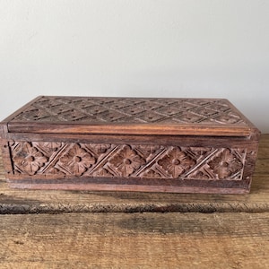 May include: A rectangular wooden box featuring a dark brown finish and intricate hand-carved floral patterns. The lid and the front panel are decorated with a repeating diamond grid design, with each section containing a carved flower motif.