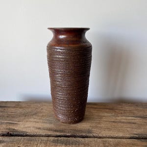 May include: A tall, cylindrical ceramic vase with a dark brown, textured finish. The exterior features horizontal ridged detailing throughout the body, leading up to a smooth, flared rim at the top. The surface has a mottled, earthy appearance.