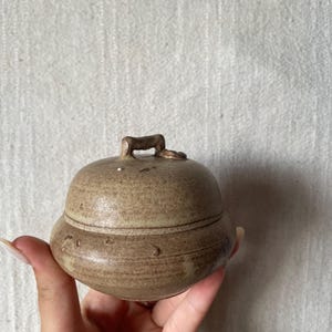 May include: A small, handmade ceramic jar with a lid. The jar is a muted brown colour with a textured surface and a small handle on top. The jar has a rounded shape and a decorative band around the middle.
