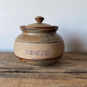 May include: A round, handcrafted ceramic sugar bowl with a lid. The vessel features a textured, tan-coloured band around the centre with the word SUGAR embossed in dark lettering. The top and bottom sections have a mottled brown and green glaze.