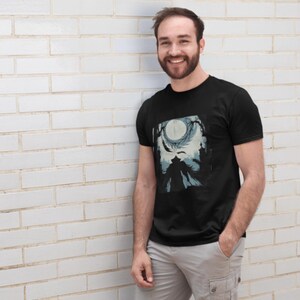 May include: Black t-shirt featuring a graphic design with a full moon, silhouette of a figure, and tree branches in shades of blue and white. The shirt is a crew neck style.
